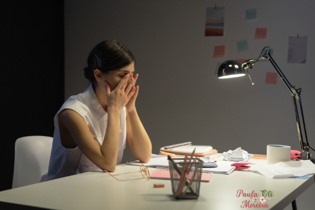 Mulher sentada em uma cadeira com cotovelos na mesa e mãos nos olhos com demostração de cansaço e estresses. Luminária monstra que é noite .