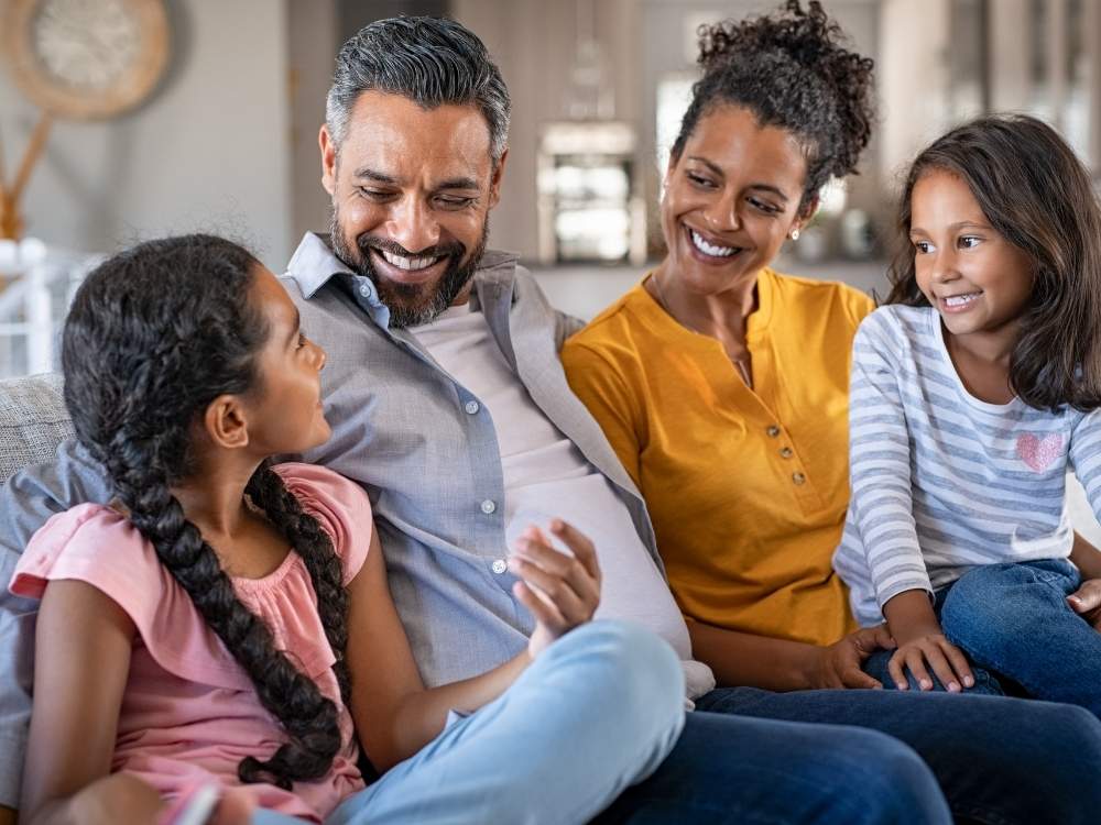 Família sentada em sofá de casa com papai, mamãe e duas filhas. Todos olham para filha maior de uns 10 anos e sorriem. 