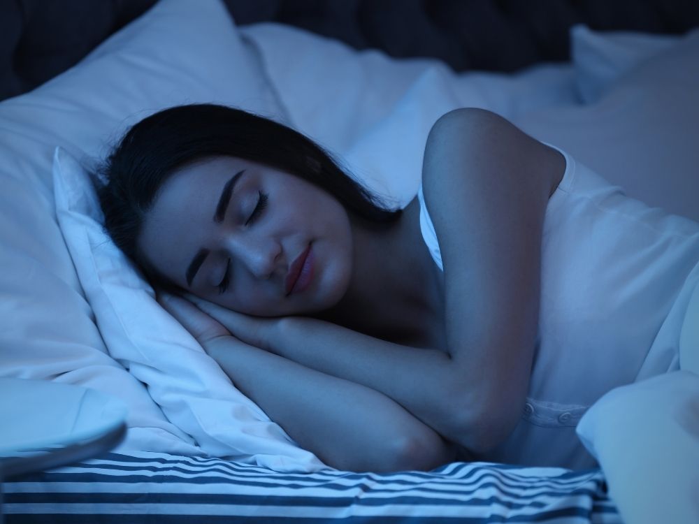 Mulher dormindo de lado em lençóis brancos, com semblante tranquilo e relaxado, sob uma luz azul suave.