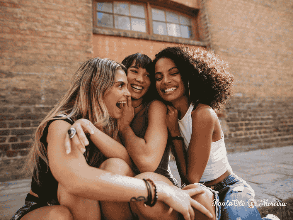 Amigas de etinias diferentes se abraçando e rindo em frente a um prédio de tijolos-Imagem:Canva